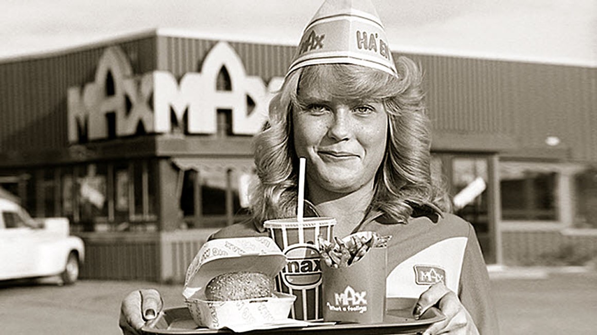 Servitris från 60-talet med MAX-kläde roch en brcika med hamburgermeny framför MAX-restaurang. Svartvit foto.