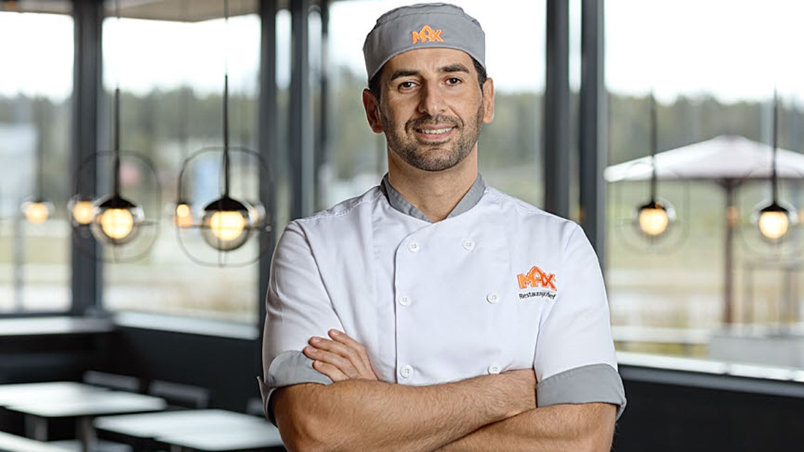 Leende manlig MAX-medarbetare i restaurangmiljö  med händerna i kors. Foto.