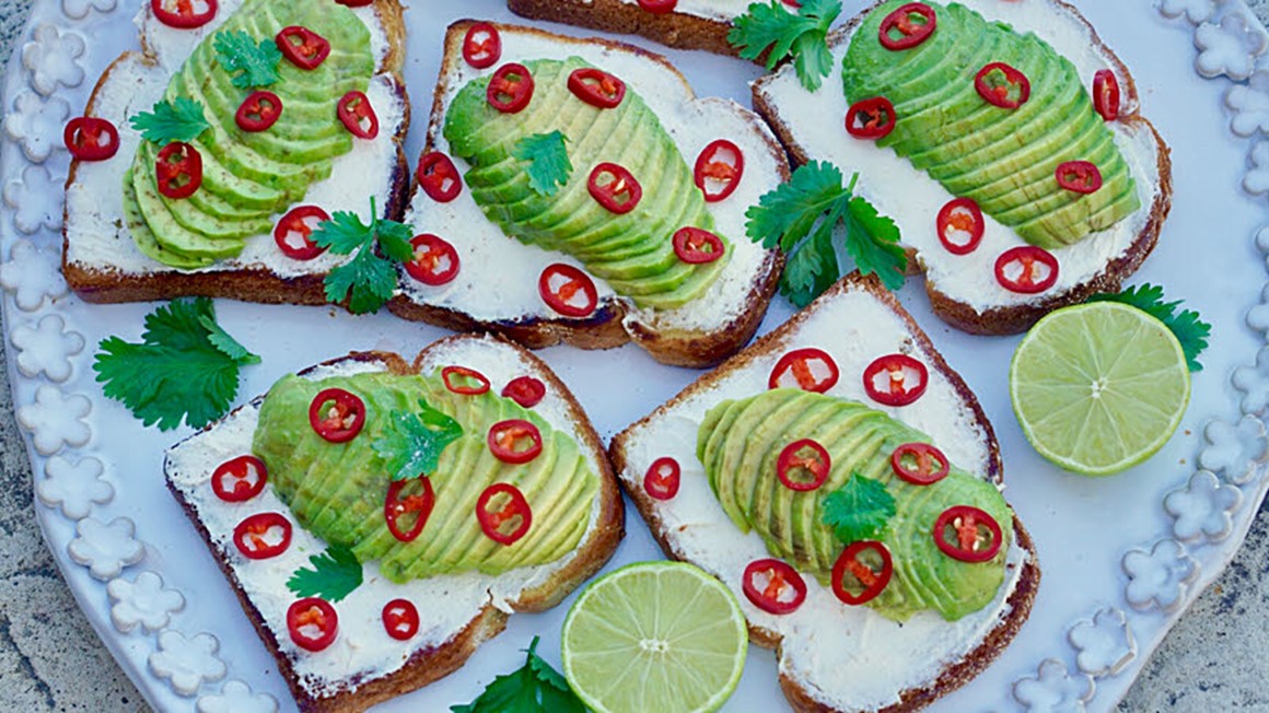 Smörgås med skivad avokado, färskost och skivad chili och persiljekvistar på stort vit fat. Foto. 
