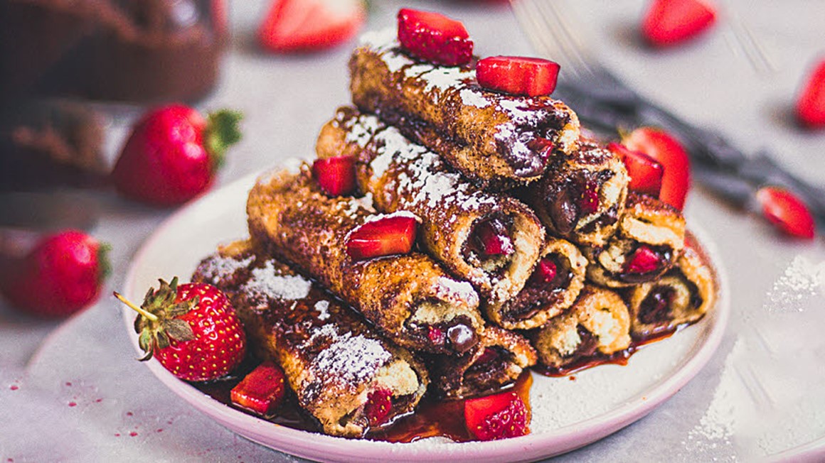 Rullade toastbröd med fyllning  av nutella och jordgubbsbitar på tallrik med hela jordgubbar i förgrunden. Foto. 