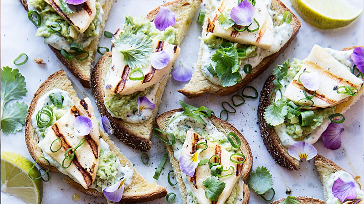 Toastsmörgås med avokadoröra, majonäs, skivad halloumi och ljust lila blomblad. Foto taget uppifrån.