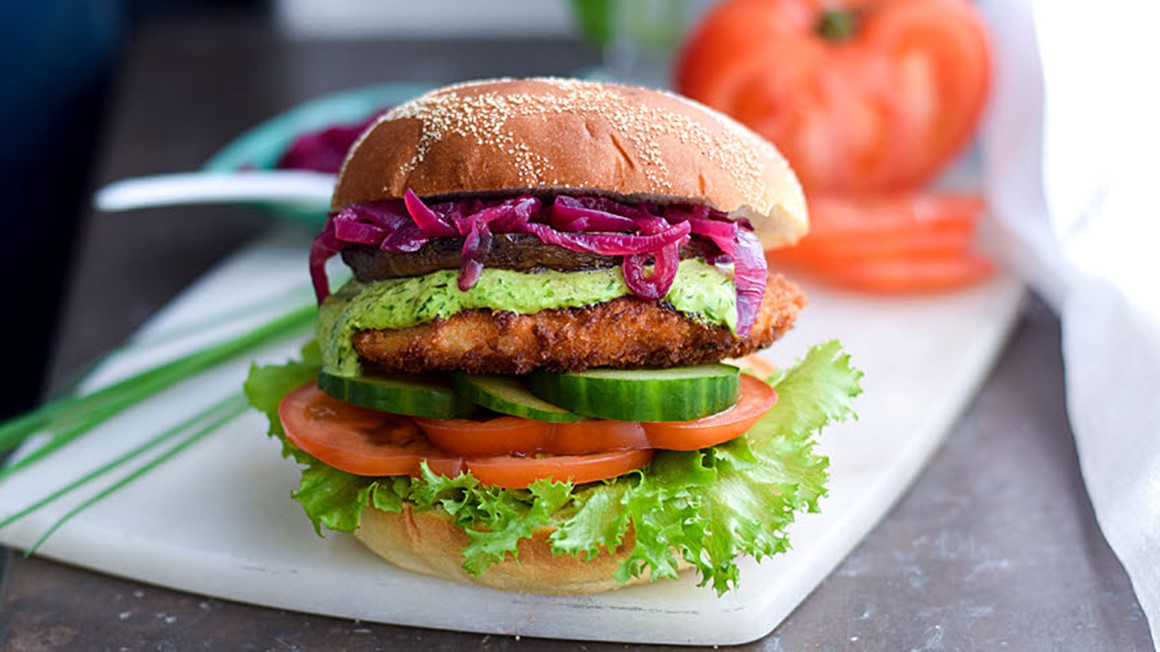 Vegetarisk hamburgare med halloumi, gurkskivor, tomatskivor och svamp på skärbräda. Foto.